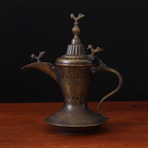 May include: An antique bronze-coloured coffee pot with a long spout and a curved handle. The pot is adorned with bird-shaped embellishments on the lid, spout, and handle. The pot is placed on a wooden surface against a dark background.