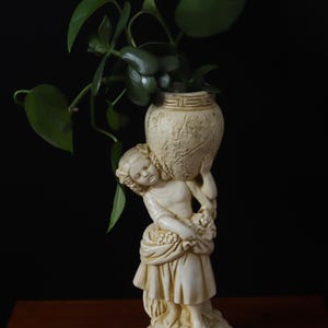 Peut inclure: Statue crème d'une jeune femme portant un vase décoratif rempli de plantes vertes à feuilles. La statue est sur une surface en bois sur fond noir. Le vase a une surface texturée.