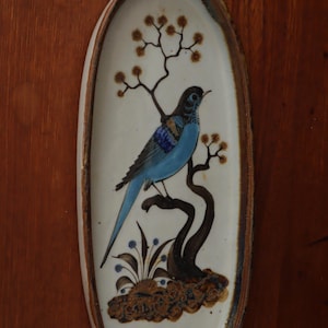 May include: Oval ceramic wall art depicting a blue bird on a brown branch with small brown flowers. The bird has blue and black details. The background is off-white with a brown border. Decorative wall art.