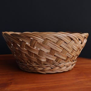 May include: A round woven basket made from light brown natural fibres. The basket has a detailed crisscross pattern and is placed on a wooden surface against a dark background. This basket is suitable for storage or display, and is measured in centimetres.