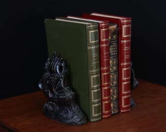 May include: A collection of books held by a black ship-shaped bookend. The books have red, green, and gold covers. The bookend is on a wooden surface against a black background.