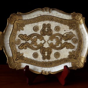 May include: A decorative serving tray, rectangular in shape, with a white and gold colour scheme. The tray has elaborate gold scrollwork and floral patterns, with handles. It is displayed on a dark surface.