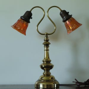 Peut inclure: Une lampe de table à deux bras en laiton avec une base et une tige en métal doré poli. Deux douilles marron foncé supportent des abat-jours en verre orange irisé, chacun orné d'un motif floral ou étoilé en relief sur la surface.