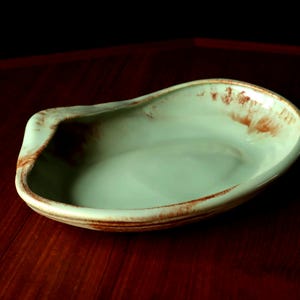 May include: A handmade, oval ceramic serving dish. The dish has a light green interior and a rim with brown accents. The dish is placed on a dark wooden surface.