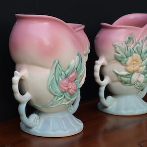 May include: Two ceramic vases featuring a soft pink gradient at the top, transitioning to a cream-coloured body and a pale blue base. Each vase is adorned with raised, sculpted floral motifs in shades of green, pink, and yellow, with handles.