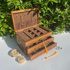 Könnte beinhalten: Eine Holzschmuckschatulle mit mehreren Schubladen und Fächern, die geöffnet ist, um verschiedene Ringe, Halsketten und Armbänder zu präsentieren. Die Schatulle hat eine braune Außenseite, einen Schlüssel und goldfarbene Beschläge. Zwei kleine Holzuntersetzer befinden sich im Vordergrund.