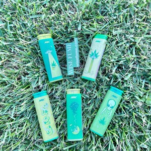 May include: Six cylindrical containers with green and yellow designs, featuring illustrations of aliens, palm trees, and the number 420. The containers are arranged on a bed of green grass, with the text "wisefume.com" visible.
