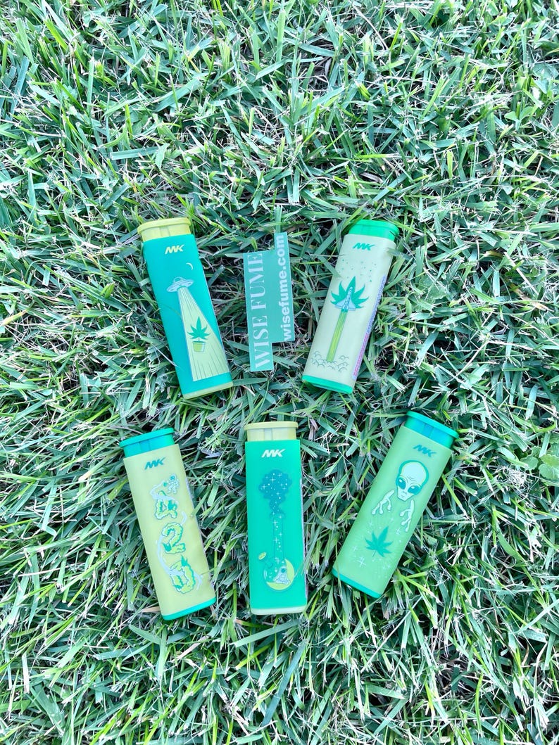 May include: Six cylindrical containers with green and yellow designs, featuring illustrations of aliens, palm trees, and the number 420. The containers are arranged on a bed of green grass, with the text "wisefume.com" visible.