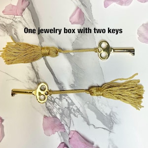 May include: Two gold-colored keys with decorative tassels, lying on a marble surface. The keys are connected to gold tassels. The text "One jewelry box with two keys" is visible.