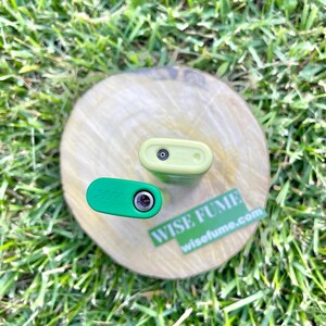 May include: Two small, rectangular, electronic devices, one green and one light green, are displayed on a wooden disc. The green device has the word "Zaza" printed on it. A sticker with the words "WISE FUME" and "wisefume.com" is also visible.
