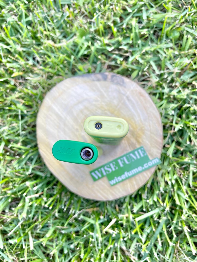 May include: Two small, rectangular, electronic devices, one green and one light green, are displayed on a wooden disc. The green device has the word "Zaza" printed on it. A sticker with the words "WISE FUME" and "wisefume.com" is also visible.