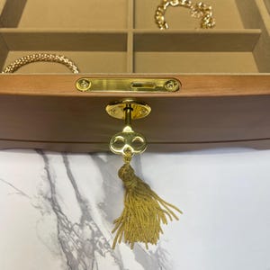 May include: A wooden jewelry box with a gold key and tassel. The interior has multiple compartments lined with tan fabric. Gold bracelets and necklaces are visible inside the box. The key is attached to the front of the box.