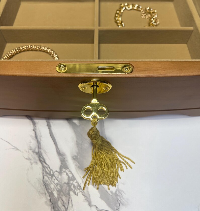 May include: A wooden jewelry box with a gold key and tassel. The interior has multiple compartments lined with tan fabric. Gold bracelets and necklaces are visible inside the box. The key is attached to the front of the box.