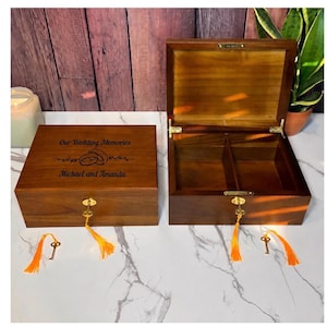 May include: Two rectangular wooden keepsake boxes with a dark brown finish. One box is closed, featuring the engraved text "Our Wedding Memories" and "Michael and Amanda" with a ring graphic. Both boxes have gold-tone locks with keys and orange tassels.