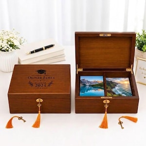 May include: Two rectangular dark wood keepsake boxes with hinged lids and gold-toned locks featuring orange tassels. One box is closed, engraved with a graduation cap and the text OLIVER JAMES CLASS OF 2024. The open box displays two landscape photos.