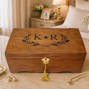 May include: A wooden jewelry box with a gold-colored tassel key. The box is brown with a decorative black floral design and the letters "K" and "R" on the lid. A small dish with jewelry and a candle are nearby.
