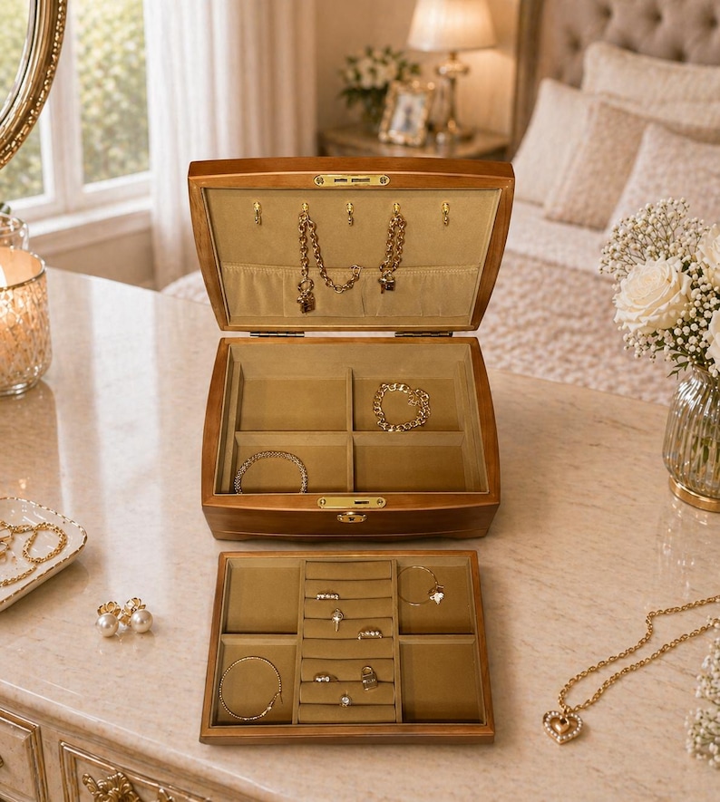 May include: A wooden jewelry box with an open lid, revealing compartments lined with beige fabric. The box contains gold necklaces, bracelets, and rings. A smaller tray with rings and bracelets sits below. The jewelry box is on a marble surface.