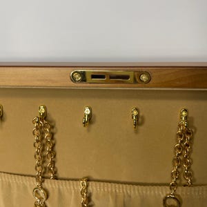May include: A jewelry box with a light brown wooden exterior and a beige interior. The interior features gold-colored hooks and chains with charms, including a key, a lock, and a cross. The box has a gold-colored clasp.