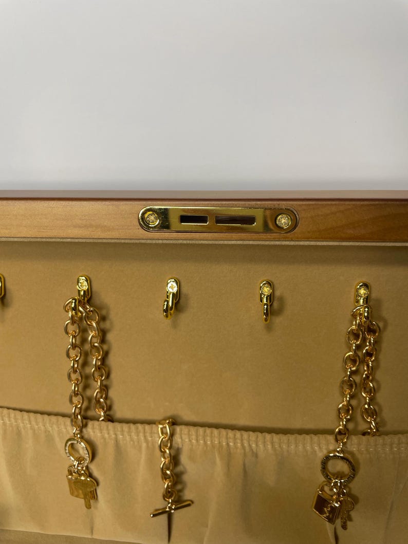 May include: A jewelry box with a light brown wooden exterior and a beige interior. The interior features gold-colored hooks and chains with charms, including a key, a lock, and a cross. The box has a gold-colored clasp.