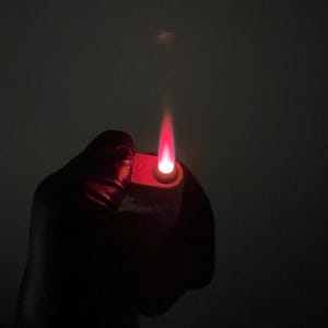 May include: A red lighter with a bright red flame against a dark background. The lighter is held in a gloved hand, with the flame extending upwards. The image is a close-up, highlighting the flame's intensity.