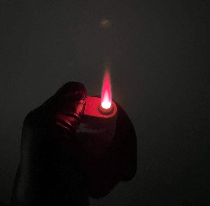 May include: A red lighter with a bright red flame against a dark background. The lighter is held in a gloved hand, with the flame extending upwards. The image is a close-up, highlighting the flame's intensity.