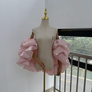 Puede incluir: Una blusa rosa claro, con hombros descubiertos y mangas abullonadas voluminosas. La prenda se exhibe en un maniquí con brazos de madera y un soporte dorado. El texto "DoodleNesta" es visible en el maniquí.