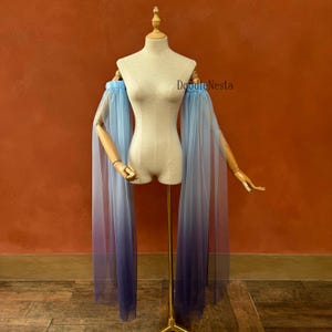 May include: A blue to purple ombre tulle sleeve cape on a mannequin. The sleeves are long and flowing, with a gathered top. The brand name "DoodleNesta" is visible on the right side of the mannequin.