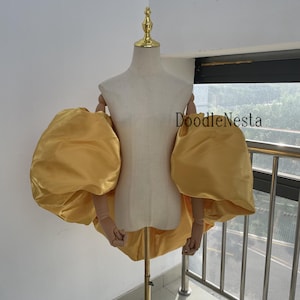 May include: A golden-yellow, voluminous sleeve garment displayed on a mannequin. The sleeves are large and puffy, made of a shiny fabric. The mannequin is neutral-toned, and the background is a light-colored wall and a window.