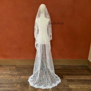 May include: A white lace wedding veil with a floral pattern, displayed on a mannequin. The veil is long, reaching the floor, and features a scalloped edge. The background is a solid reddish-brown color.