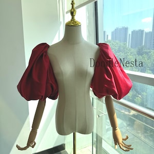 Puede incluir: Una blusa roja con mangas abullonadas exhibida en un maniquí. Las mangas son voluminosas y la tela parece ser satén o similar. El maniquí es de color crema, y el fondo incluye una ventana con vista a la ciudad. El texto "DoodleNesta" es visible.