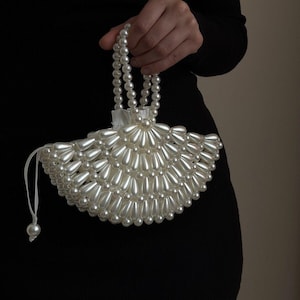 May include: A white, fan-shaped handbag made of pearlescent beads. The bag has a beaded handle and a drawstring closure with a single bead. The bag is held against a dark background.
