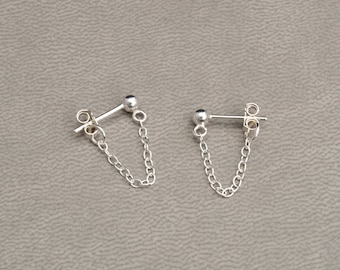 May include: A pair of silver stud earrings with a small ball on the front and a chain that dangles down to the back. The earrings are displayed on a gray surface.