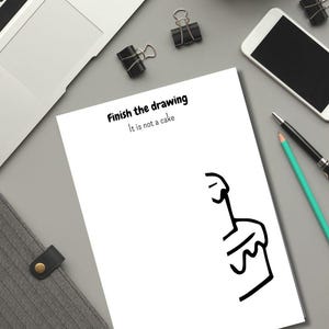 May include: A white sheet of paper with the text "Finish the drawing" and "It is not a cake" in black. A partially drawn line art image is on the right side. A laptop, phone, and office supplies are also visible.