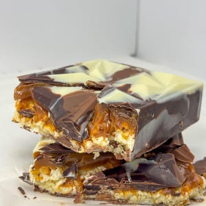 May include: Three chocolate caramel bars stacked on top of each other. The bars feature a biscuit base, a caramel filling, and a chocolate topping. The chocolate is dark and white, with a geometric pattern.