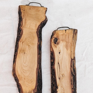 Può includere: Due vassoi di legno da portata con bordi naturali e manici in metallo. I vassoi sono realizzati in legno chiaro con bordi di corteccia marrone scuro. La venatura del legno è visibile e i vassoi sono rettangolari.