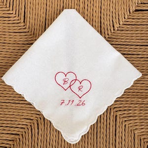May include: A white square handkerchief with scalloped edges featuring red embroidery of two interlocking hearts. Inside the hearts are the letters B and R, and below the hearts is the embroidered date 7.11.26 in a matching red thread.