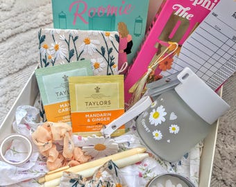 Op de afbeelding: Een geschenkdoos gevuld met diverse items, waaronder boeken, thee, kaarsen en accessoires. De doos bevat een boek getiteld "The Temporary Roomie", een kaars en een waterfles met madeliefjesontwerpen.