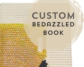 May include: A custom bedazzled book with a white background and a section covered in sparkling yellow and gold rhinestones. The text "CUSTOM BEDAZZLED BOOK" is displayed in black letters on a beige circular background.