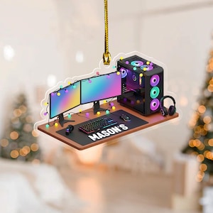 May include: A gaming-themed ornament featuring a computer setup with two monitors, a keyboard, mouse, and headset. The desk mat has the name "MASON'S" printed on it. The computer case has colorful lights. The ornament is suspended by a gold string.