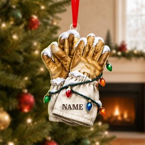 May include: A Christmas tree ornament shaped like a pair of tan leather work gloves covered in patches of white snow. The gloves are wrapped in a string of colorful Christmas lights and feature the word NAME printed on the cuff.