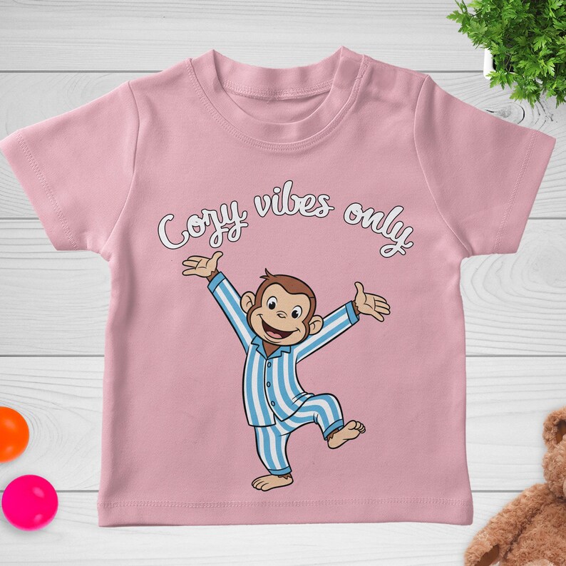 Op de afbeelding: Roze kindershirt met de tekst "Cozy Vibes Only" in witte letters. Het shirt heeft een cartoon aap in een blauw-wit gestreepte pyjama met uitgestrekte armen. Een leuk en comfortabel ontwerp voor kinderen.