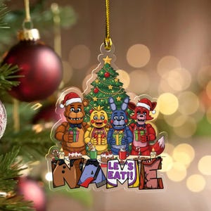 May include: A Christmas ornament featuring cartoon characters wearing Santa hats, holding gifts, and standing in front of a decorated Christmas tree. The ornament has the words "NAME" and "LET'S EAT!!" at the bottom. The ornament is hanging from a gold string.