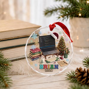 May include: A round clear acrylic ornament featuring a laptop with a Santa hat, a stack of books labeled PLAN, CODE, DEBUG, DEPLOY, REPEAT, a mug with text EAT CODE REPEAT, a keyboard, a Christmas tree, and the name BRIAN 2026 with snowflakes.