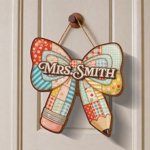 May include: A wooden sign shaped like a bow and pencil, with the text "MRS. SMITH" in a brown, decorative font. The bow and pencil are made of patchwork fabric patterns in various colors, including red, blue, and yellow. The sign hangs from a wooden peg.