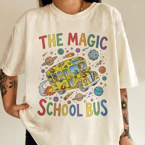 May include: Cream-colored t-shirt featuring a colorful graphic of a yellow school bus in space, with the text "THE MAGIC SCHOOL BUS". The bus has passengers and is surrounded by planets, stars, and comets. A fun, whimsical design.