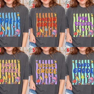 May include: Dark gray t-shirts with colorful grade level designs. Each shirt features a grade level like "Pre-K" or "Kindergarten" in a vertical striped design with school-themed graphics, including pencils, apples, and backpacks. The text "Mrs. Alice" is at the bottom.