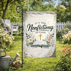 May include: A garden flag with the text "It's Too Nonfictiony Outside" in dark green script. The flag features a gold cross, an open book, and floral accents on a cream background. The flag is displayed on a black metal stand in a garden setting.