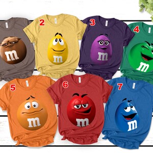 May include: A collection of seven short-sleeved t-shirts in various colors, each featuring a different M&M's character graphic. The shirts are brown, yellow, purple, green, orange, red, and blue. Each graphic includes the letter "m".