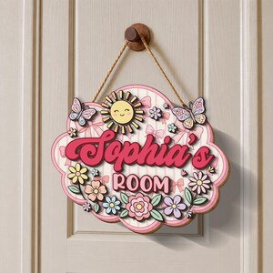 May include: A wooden sign for a room, featuring the name "Sophia's ROOM" in a pink, cursive font. The sign is decorated with a smiling sun, butterflies, flowers, and bows. It hangs from a wooden knob with a rope.