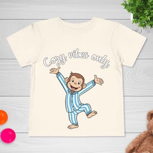 Op de afbeelding: Cr&egrave;mekleurig T-shirt met een cartoon aap in blauw-witte gestreepte pyjama met uitgestrekte armen. De tekst "Cozy vibes only" staat in een speels lettertype boven de aap. Een leuk en comfortabel T-shirt voor kinderen.